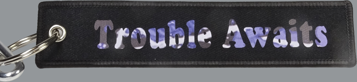 A rectangular black fabric key tag displayed against a neutral gray background. The tag has a textured woven surface with stitched edges and a metal grommet on the left side, attached to a silver keyring. Centered on the tag is the phrase “Trouble Awaits” in a bold, clean font. The lettering features a blue and purple camo-style pattern with subtle color variation, giving it a slightly reflective and textured appearance. 