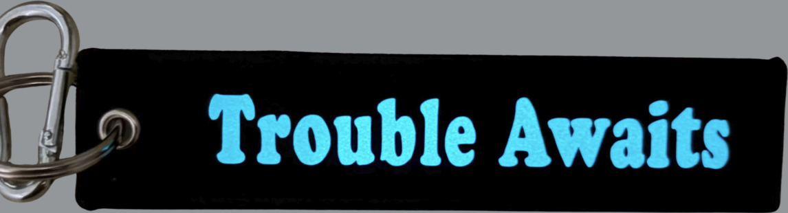 A rectangular black fabric key tag displayed against a neutral gray background. The tag has a textured woven surface with stitched edges and a metal grommet on the left side, attached to a silver keyring and clip. Centered on the tag is the phrase “Trouble Awaits” in a bold, rounded font. The lettering appears in a bright blue color with a glow-in-the-dark effect, standing out strongly against the black fabric. The overall design combines a durable textile base with a high-contrast, luminous logo 