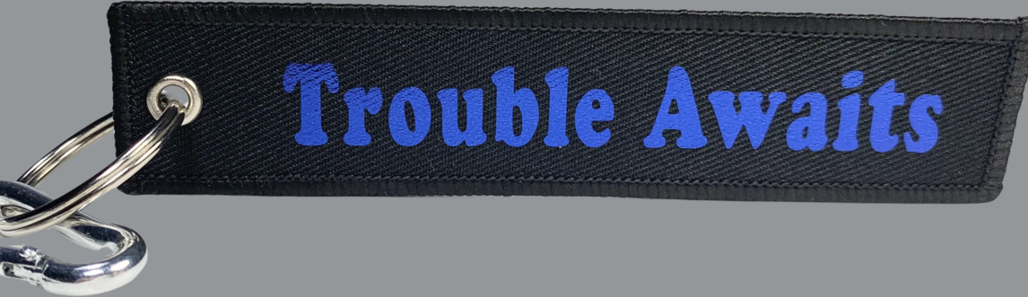 A rectangular black fabric key tag displayed against a neutral gray background. The tag has a textured woven surface with stitched edges and a metal grommet on the left side, attached to a silver keyring and clip. Centered on the tag is the phrase “Trouble Awaits” in a bold, rounded font. The lettering appears in a metallic blue finish, giving it a slightly reflective look that contrasts against the matte black fabric. The overall design combines a durable textile base with a clean, high-contrast logo 