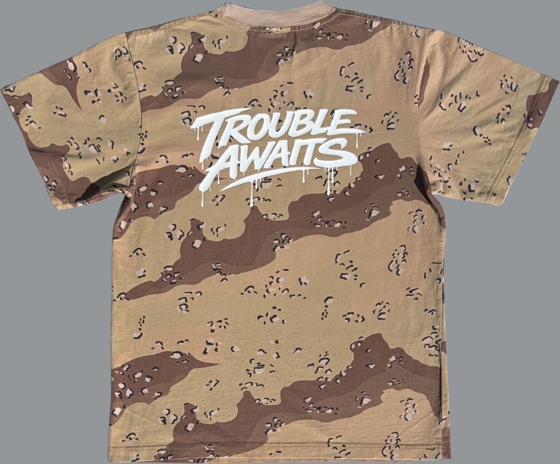 A back view of an oversized, boxy short-sleeve T-shirt displayed flat against a neutral gray background. The shirt features a desert camouflage pattern made up of light tan, sandy beige, and medium brown shapes with small scattered darker markings. Centered across the upper back is a large, bold graphic reading “Trouble Awaits” in a stylized graffiti-inspired script. The lettering is white with a slightly glossy appearance and includes dripping paint details extending downward from several letters.
