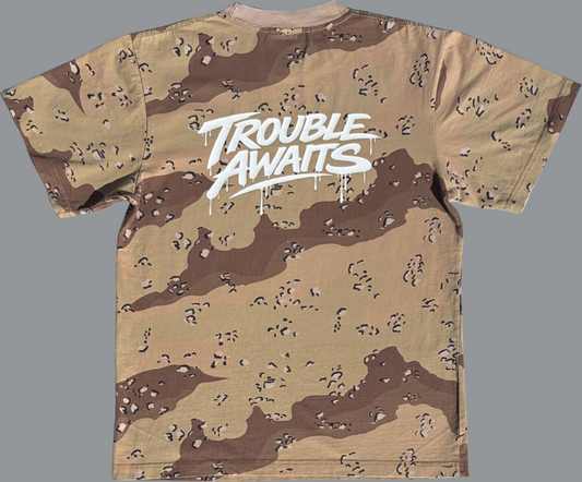 A back view of an oversized, boxy short-sleeve T-shirt displayed flat against a neutral gray background. The shirt features a desert camouflage pattern made up of light tan, sandy beige, and medium brown shapes with small scattered darker markings. Centered across the upper back is a large, bold graphic reading “Trouble Awaits” in a stylized graffiti-inspired script. The lettering is white with a slightly glossy appearance and includes dripping paint details extending downward from several letters.
