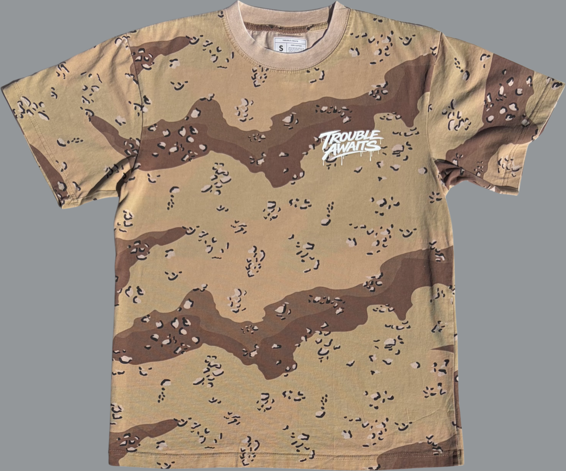 A front view of an oversized, boxy short-sleeve T-shirt displayed flat against a neutral gray background. The shirt features a desert camouflage pattern composed of sandy beige, light tan, and medium brown shapes, with small scattered darker speckles throughout.

On the upper left chest, there is a small white graphic that reads “Trouble Awaits” in a graffiti-style script with subtle dripping paint details beneath the lettering. The print contrasts sharply against the camo background