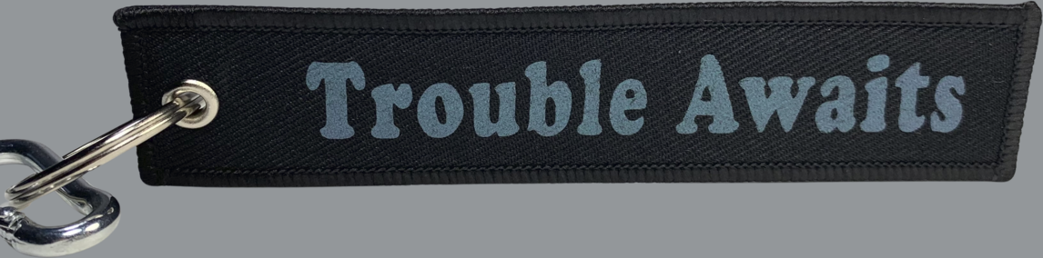A rectangular black fabric key tag displayed against a neutral gray background. The tag has a textured woven surface with stitched edges and a metal grommet on the left side, attached to a silver keyring and clip. Centered on the tag is the phrase “Trouble Awaits” in a bold, rounded font. The lettering features a subtle color-shifting finish that appears gray at first but reflects faint tones of green and purple depending on the angle and lighting. The overall design is understated
