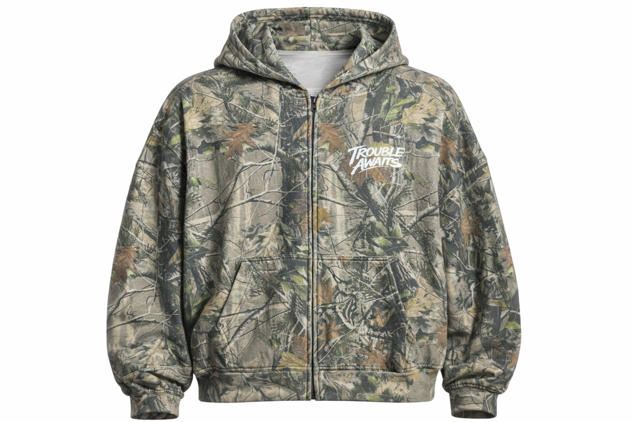 Front-facing ghost mannequin image of an oversized boxy zip-up hoodie with dropped shoulders in a woodland tree camouflage print. The hoodie features a full front zipper, roomy hood, ribbed cuffs and hem, kangaroo-style front pockets, and a white “Trouble Awaits” logo printed on the left chest.
