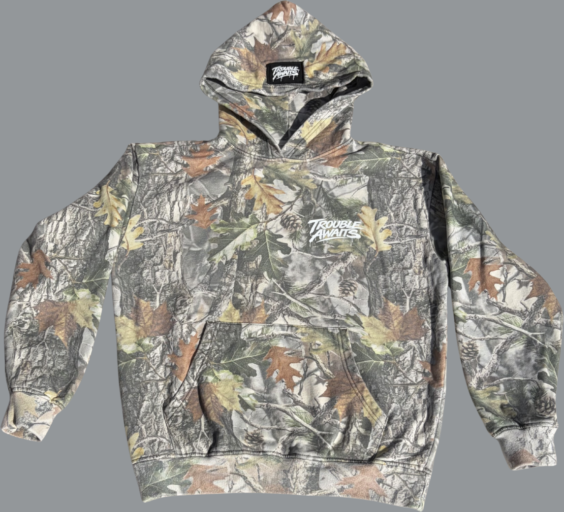 This image shows the front view of a pullover hoodie laid flat against a neutral gray background. The hoodie features a detailed forest camouflage pattern composed of tree bark textures, branches, and scattered leaves in shades of green, brown, tan, and gray. On the left chest area, there is a small white “Trouble Awaits” logo printed in a graffiti-inspired script with subtle paint-drip accents. The hoodie includes a front kangaroo pocket, a hood with a visible interior label at the neckline, long sleeves