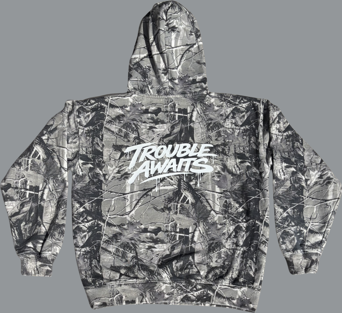 This image shows the back view of a pullover hoodie laid flat against a neutral gray background. The hoodie features a grayscale camouflage pattern composed of branches, leaves, and natural textures in varying shades of gray, black, and muted tones. Across the upper back is a large white “Trouble Awaits” logo printed in a bold, graffiti-inspired script with paint-drip effects extending downward from the lettering. The hood is visible at the top, and the garment has long sleeves with ribbed cuffs