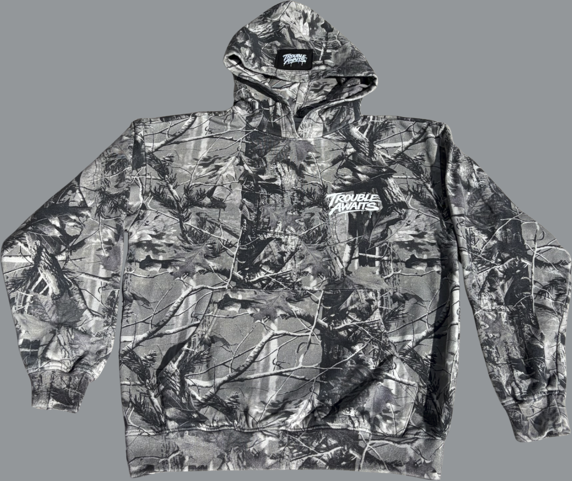 This image shows the front view of a pullover hoodie laid flat against a neutral gray background. The hoodie features a grayscale camouflage pattern composed of branches, leaves, and natural textures in shades of gray, black, and muted tones. On the left chest area, there is a small white “Trouble Awaits” logo printed in a graffiti-inspired script with subtle paint-drip accents. The hoodie includes a front kangaroo pocket, a hood with a visible interior label at the neckline, long sleeves, and ribbed cuffs 