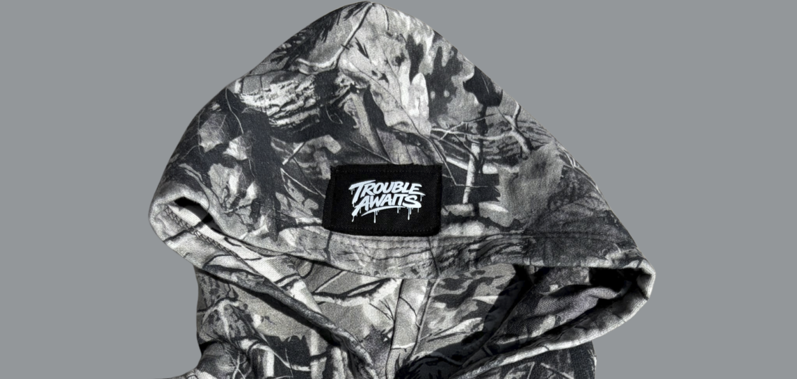 This image shows a close-up of the hood area of a grayscale camouflage hoodie. The fabric features a detailed monochrome camo pattern made up of branches, leaves, and natural textures in varying shades of gray and black. Centered on the front edge of the hood is a small rectangular black woven patch displaying the “Trouble Awaits” logo in white, styled in a graffiti-inspired script with subtle drip details. The hood is slightly open, revealing a darker interior lining. 