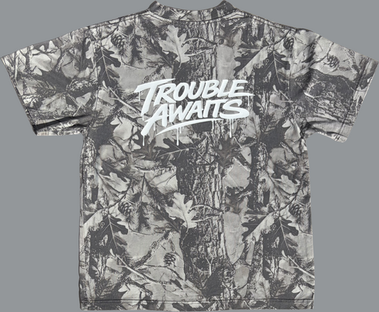 A short-sleeve T-shirt is displayed flat against a plain gray background, shown from the back. The shirt has an oversized, boxy fit with slightly wide sleeves and a straight hem.

The fabric features a monochrome woodland camouflage pattern in shades of gray, black, and off-white. The design includes detailed elements such as tree bark textures, branches, leaves, and pinecones layered across the entire surface, creating a realistic forest-inspired print.


