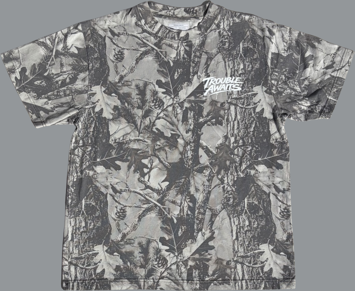 A short-sleeve T-shirt is displayed flat against a plain gray background, shown from the front. The shirt has an oversized, boxy fit with wide sleeves, a crew neckline, and a straight hem.

The fabric features a monochrome woodland camouflage pattern in shades of gray, black, and off-white. The print includes detailed elements such as tree bark textures, branches, leaves, and pinecones layered across the entire surface, creating a realistic forest-inspired design.

