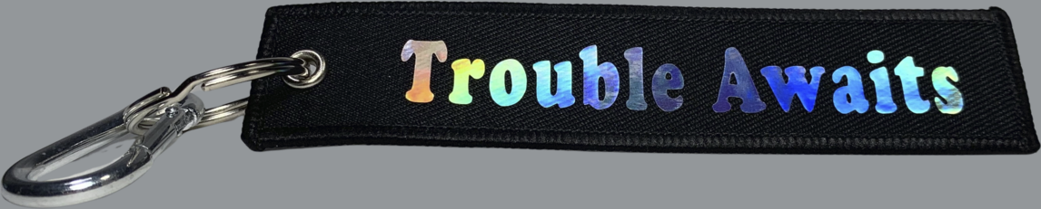 A rectangular black fabric key tag displayed against a neutral gray background. The tag has a textured woven surface with stitched edges and a metal grommet on the left side, attached to a silver keyring and clip. Centered on the tag is the phrase “Trouble Awaits” in a bold, rounded font. The lettering features a holographic finish that reflects shifting rainbow tones, including blue, green, purple, and hints of yellow depending on the light. The reflective text contrasts with the matte black fabric, 