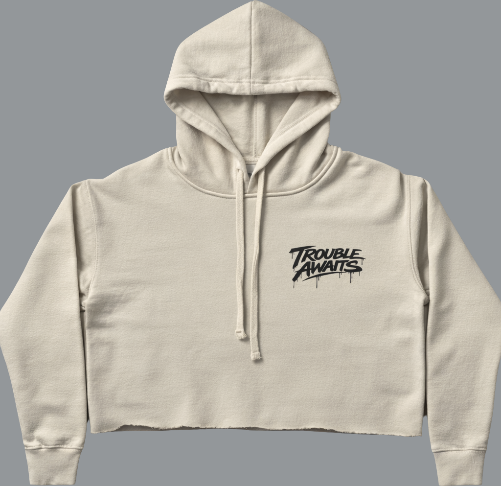 Front view of a women’s cropped hoodie in a light cream/beige color. The hoodie has a relaxed, slightly oversized fit with dropped shoulders and long sleeves that taper into ribbed cuffs. It features a drawstring hood with matching cream cords hanging at the center. The hem is raw-cut, creating a casual, unfinished edge. On the upper left chest is a small black “Trouble Awaits” logo in a graffiti-style script with subtle paint-drip details. The garment is displayed against a plain neutral gray background.