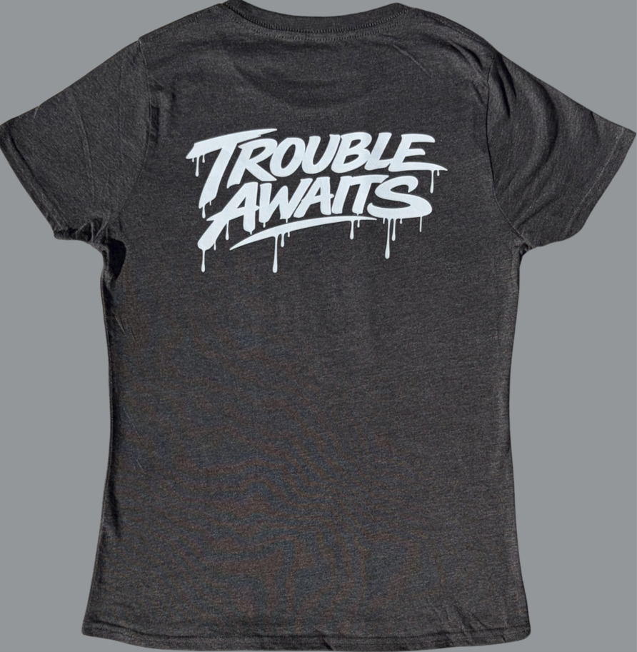 A back view of a women’s fitted T-shirt in a heather gray fabric, displayed against a neutral gray background. The shirt has a slightly contoured, tapered shape with short sleeves and a crew neckline.

Across the upper back is a bold white “Trouble Awaits” graphic in a graffiti-inspired font with dripping paint details, giving it a streetwear aesthetic. The design spans the shoulder blade area and contrasts strongly against the darker gray fabric.

The material appears soft and lightweight