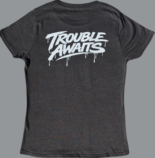 A back view of a women’s fitted T-shirt in a heather gray fabric, displayed against a neutral gray background. The shirt has a slightly contoured, tapered shape with short sleeves and a crew neckline.

Across the upper back is a bold white “Trouble Awaits” graphic in a graffiti-inspired font with dripping paint details, giving it a streetwear aesthetic. The design spans the shoulder blade area and contrasts strongly against the darker gray fabric.

The material appears soft and lightweight