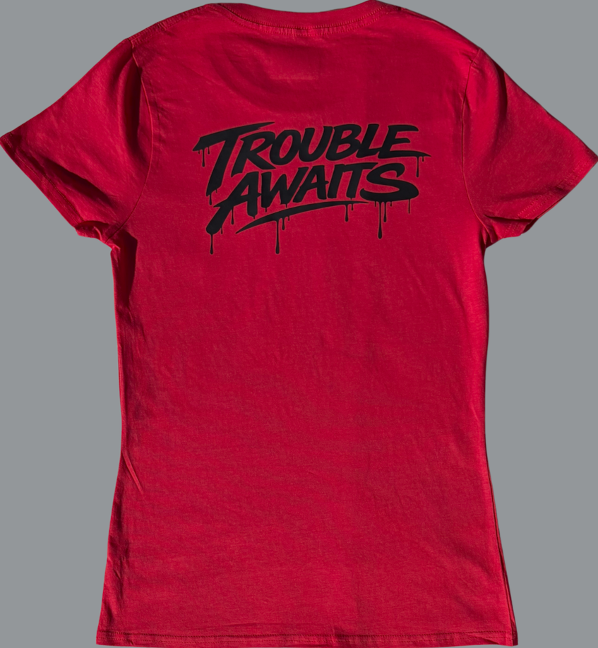 A back view of a women’s lightweight T-shirt in a rich red color, displayed against a neutral gray background. The shirt has a slightly fitted, tapered shape with short sleeves and a crew neckline.

Centered across the upper back is a bold black graphic reading “Trouble Awaits” in a stylized graffiti-inspired font with dripping paint details, giving it a streetwear aesthetic. The print sits across the shoulder blade area and contrasts sharply against the red fabric.

The fabric appears soft and lightweight 