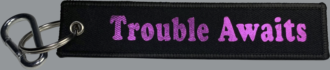 A rectangular black fabric key tag displayed against a neutral gray background. The tag has a textured woven surface with stitched edges and a metal grommet on the left side, attached to a silver keyring and clip. Centered on the tag is the phrase “Trouble Awaits” in a bold, rounded font. The lettering appears in a metallic magenta or pink finish with a slightly reflective, glossy look, creating a vibrant contrast against the black fabric. 