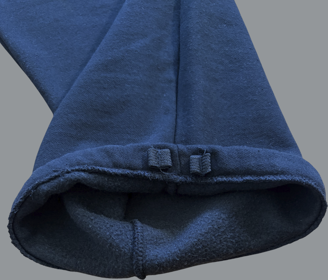 Close-up of the bottom hem of black sweatpants shown turned inside out. The image highlights the straight-leg opening with no elastic cuff, exposing the soft fleece interior lining. Visible stitching runs along the hem, and two small fabric tabs or loops are sewn near the edge. The material appears thick and textured, emphasizing a heavyweight, cozy construction.