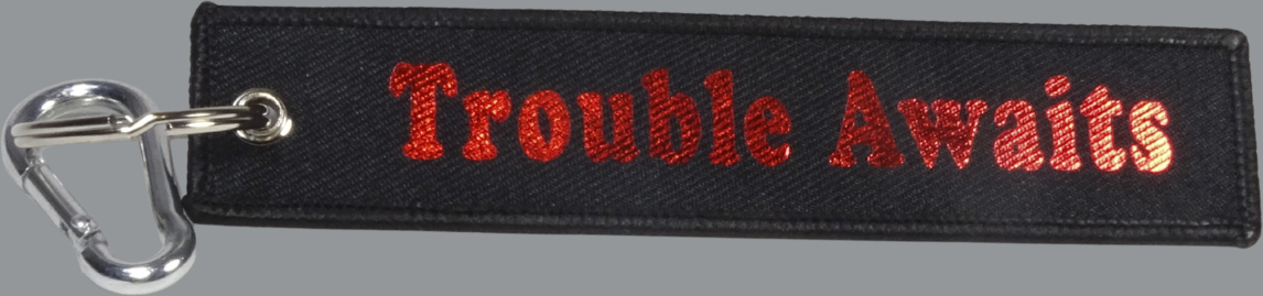 A rectangular black fabric key tag displayed against a neutral gray background. The tag has a textured woven surface with stitched edges and a metal grommet on the left side, attached to a silver keyring and clip. Centered on the tag is the phrase “Trouble Awaits” in a bold, rounded font. The lettering appears in a metallic red finish with a slightly reflective, glossy look, standing out vividly against the black fabric. 
