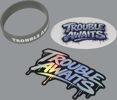 A product set displayed on a neutral gray background featuring three “Trouble Awaits” items. In the top left is a matte gray silicone wristband with bold white uppercase text reading “TROUBLE AWAITS” printed around the band. In the top right is an oval-shaped sticker with a graffiti-style “Trouble Awaits” design in a blue and purple gradient, surrounded by a dark spray-paint outline with splatter and drip effects. At the bottom is a die-cut holographic sticker featuring the same “Trouble Awaits” 
