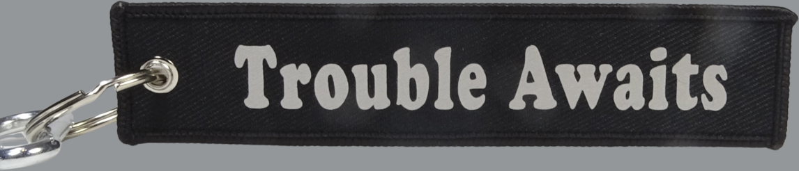 A rectangular black fabric key tag displayed against a neutral gray background. The tag has a textured woven surface with stitched edges and a metal grommet on the left side, attached to a silver keyring and clip. Centered on the tag is the phrase “Trouble Awaits” in a bold, rounded font. The lettering appears in a solid light silver or white tone, creating a clean, high-contrast look against the black fabric. 