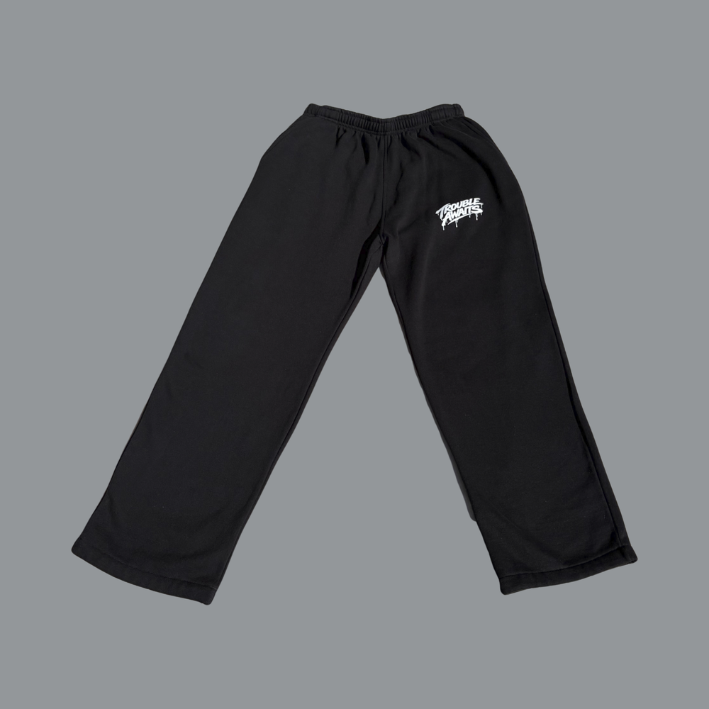 Black baggy sweatpants laid flat against a neutral gray background. The pants feature a relaxed, straight-leg fit with no cuffs, a gathered elastic waistband, and wide leg openings. A small white “Trouble Awaits” logo is printed on the upper right thigh. The fabric appears soft and slightly structured, with subtle folds along the legs indicating a loose, oversized silhouette.