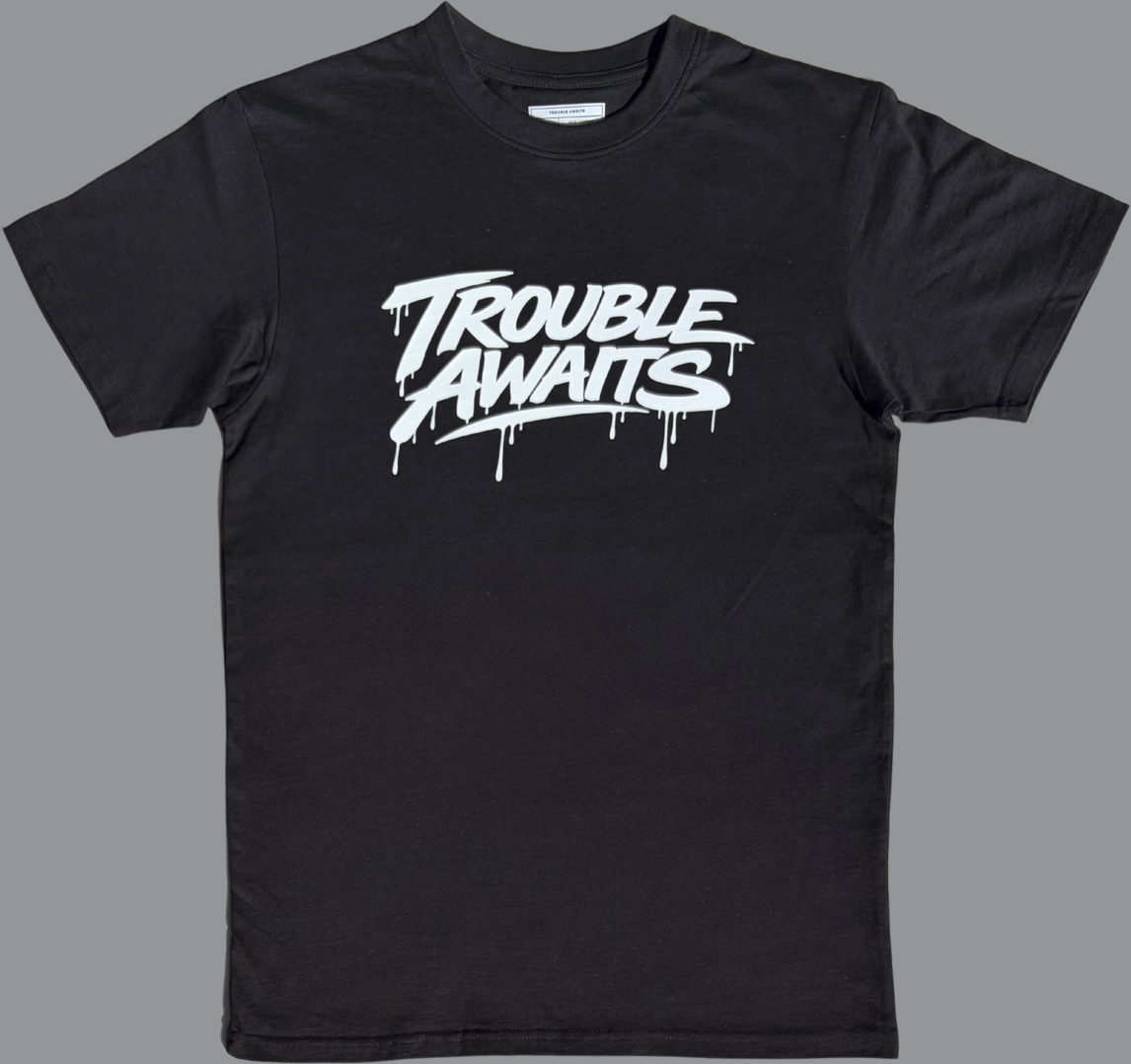 Black short sleeve t-shirt featuring a large off white graphic centered on the chest. The graphic reads “Trouble Awaits” in bold stylized lettering with a dripping paint effect beneath several of the letters. The design has a graffiti-inspired look that contrasts sharply against the black fabric. The rest of the shirt is plain black with no additional graphics or patterns visible on the front.