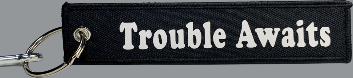 A rectangular black fabric key tag displayed against a neutral gray background. The tag has a textured woven surface with stitched edges and a metal grommet on the left side, attached to a silver keyring and clip. Centered on the tag is the phrase “Trouble Awaits” in a bold, rounded font. The lettering appears in solid white, creating strong contrast against the black fabric for clear readability. The overall design is simple and clean, combining a durable textile base with a highly legible logo 