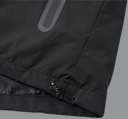 A detailed close-up of the lower corner of a black windbreaker jacket shows the hem construction. The fabric appears smooth and lightweight with a slight sheen. An adjustable drawcord runs through the hem, exiting at the side and secured with a small black plastic toggle for tightening. Subtle stitching lines and gentle fabric creases are visible, indicating a flexible, water-resistant material.