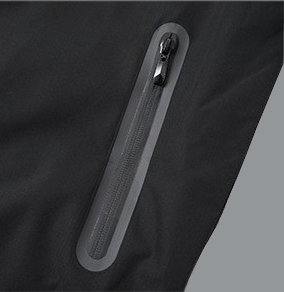 A close-up detail of a black windbreaker shows a vertical zippered pocket. The zipper is coated and outlined with a smooth, slightly lighter gray waterproof seal, giving it a sleek, technical look. A small black zipper pull is attached at the top. The surrounding fabric appears lightweight with a subtle matte sheen and fine texture, consistent with water-resistant outerwear.