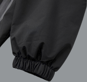 A detailed close-up of a black windbreaker sleeve shows an elasticated cuff. The fabric gathers tightly at the wrist, creating a slightly puffed look above the cuff. The material appears lightweight with a subtle sheen and soft creasing, consistent with water-resistant outerwear. Clean stitching lines are visible, emphasizing a functional, streamlined design.