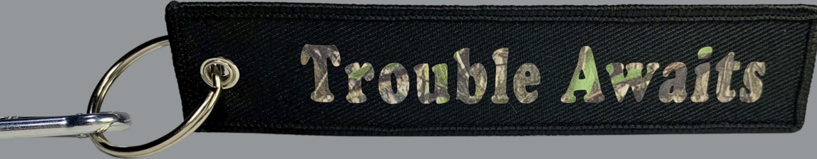 A rectangular black fabric key tag displayed against a neutral gray background. The tag has a textured woven surface with stitched edges and a metal grommet on the left side, attached to a silver keyring and clip. Centered on the tag is the phrase “Trouble Awaits” in a bold, rounded font. The lettering features a woodland camouflage pattern with mixed tones of green, brown, and tan, giving it a rugged, outdoor-inspired look. The camo text contrasts subtly against the black fabric